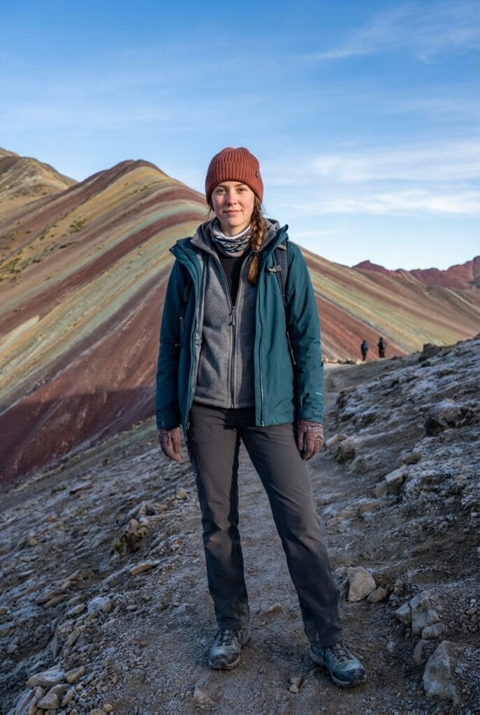 A woman wearing outdoor clothing stands on a rocky trail with Rainbow Mountain’s colorful striped slopes in the background under a clear sky.