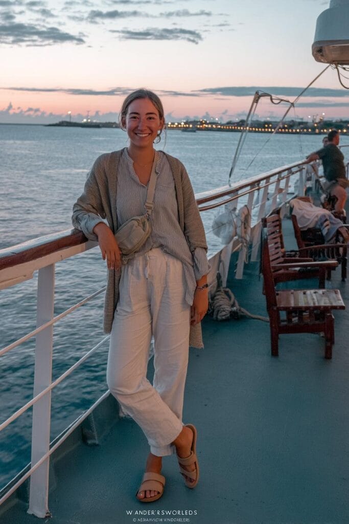 A woman smiles while standing on the deck of a boat at sunset, with water and city lights visible in the background.