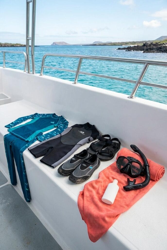 Snorkeling gear, swimwear, sandals, and a towel are laid out on a boat seat, with clear blue ocean and rocky islands visible in the background.